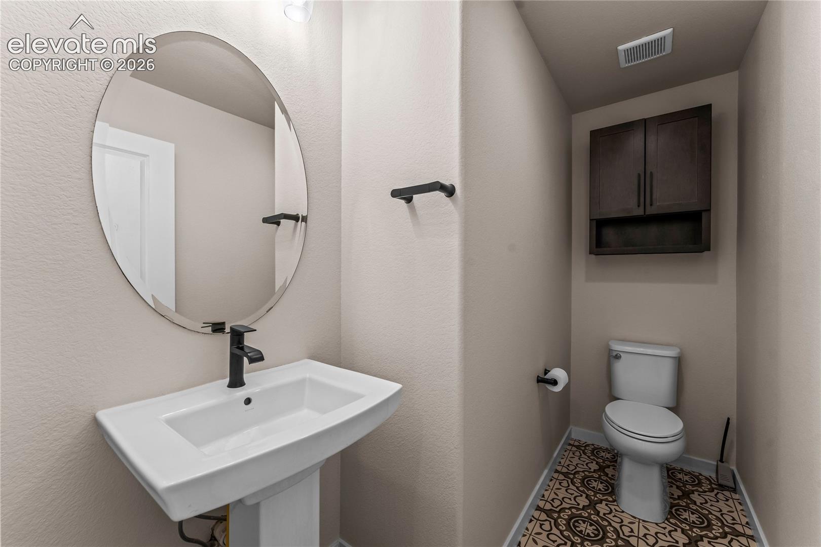 Pedestal sink with matte black faucet, oval wall-mounted mirror, decorative patterned tile flooring, and dark wood-finish wall cabinetry