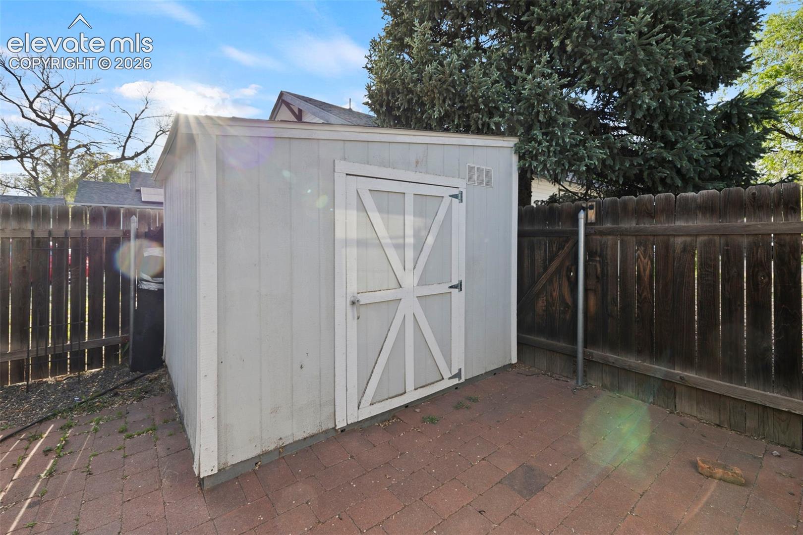 Storage shed with a barn-style door, set on a brick paver patio, featuring a wood fence perimeter