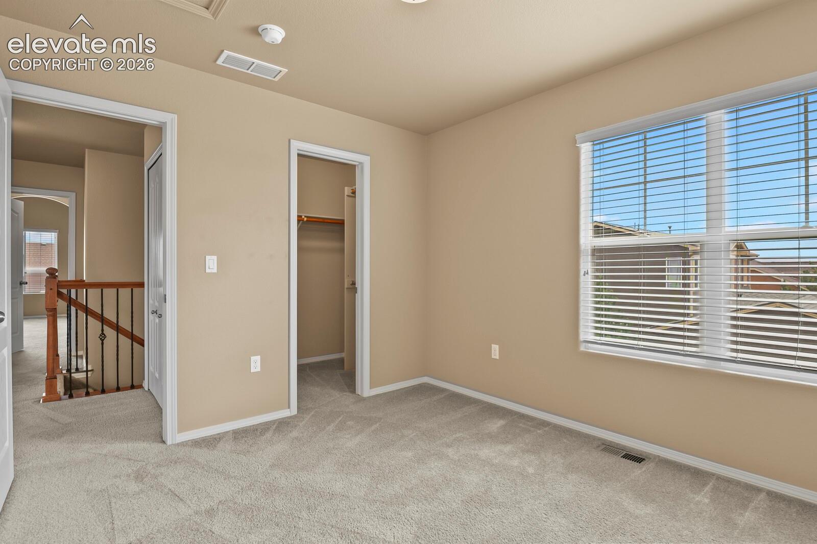 Carpeted flooring, neutral wall tones, window with horizontal blinds, open closet with hanging rod, and a staircase with wood and metal railing