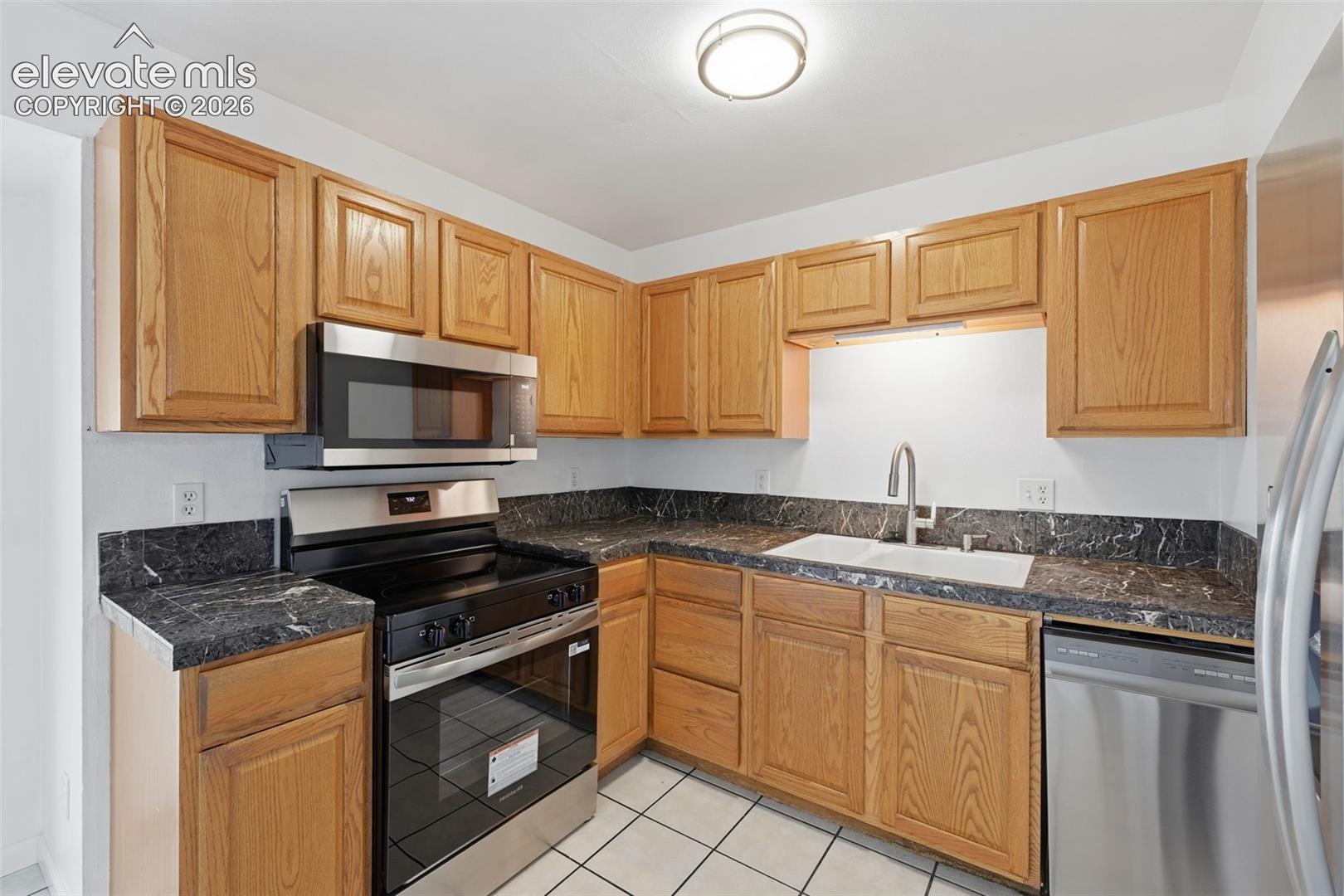 Kitchen featuring wood cabinetry, dark countertops, white tile flooring, stainless steel appliances, and a double basin sink