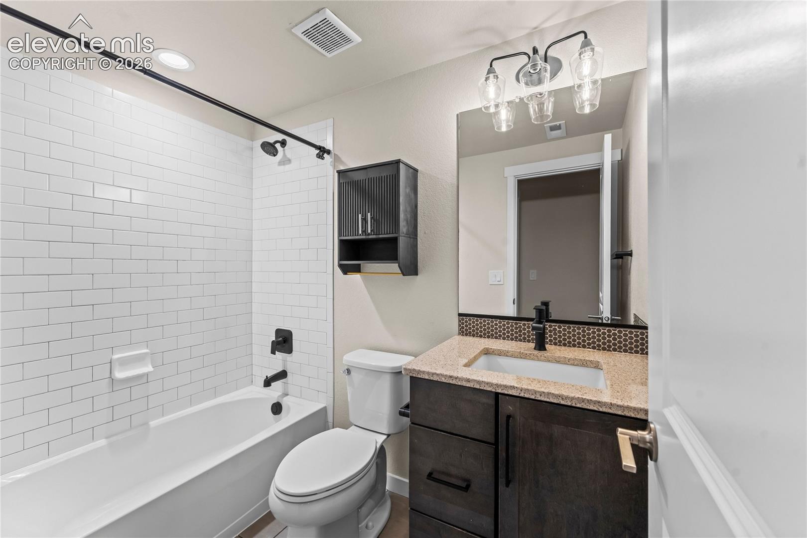 Bathroom featuring a tub-shower combination with white subway tile surround, vanity with stone-finish countertop and integrated sink, matte black plumbing fixtures, and wall-mounted storage cabinet