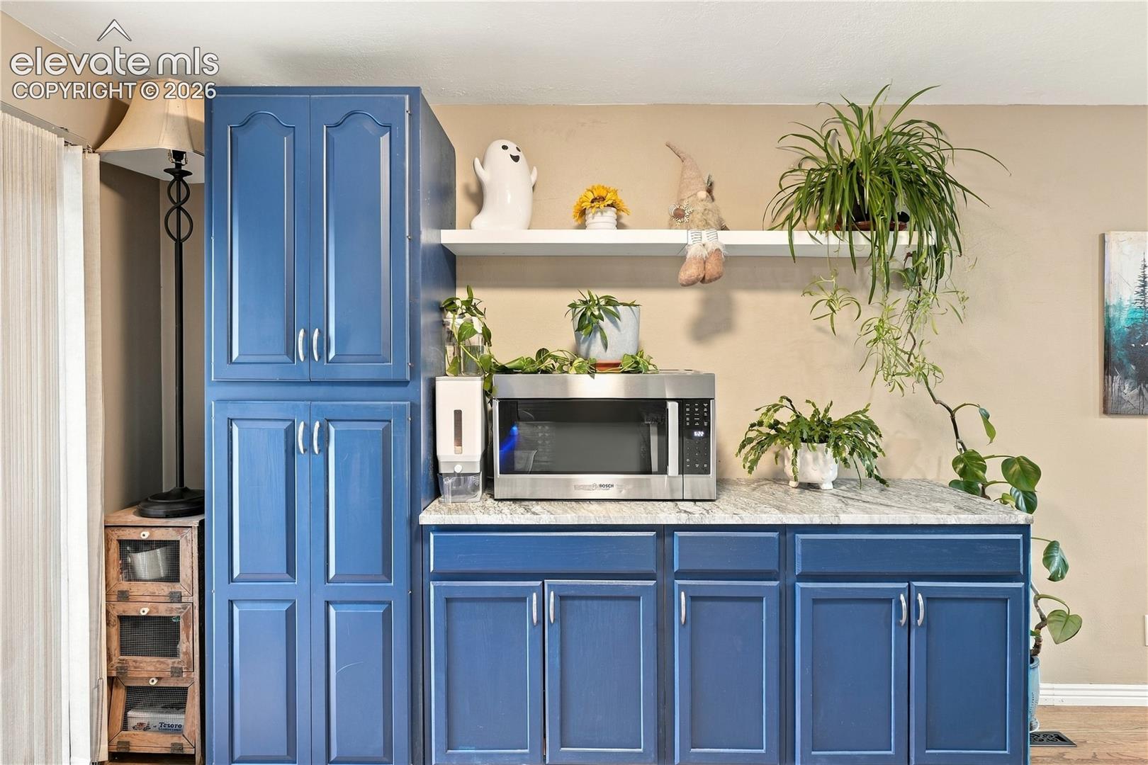 Blue-painted cabinetry featuring raised-panel doors, stone-finish countertop, integrated stainless steel microwave, and wall-mounted white shelving