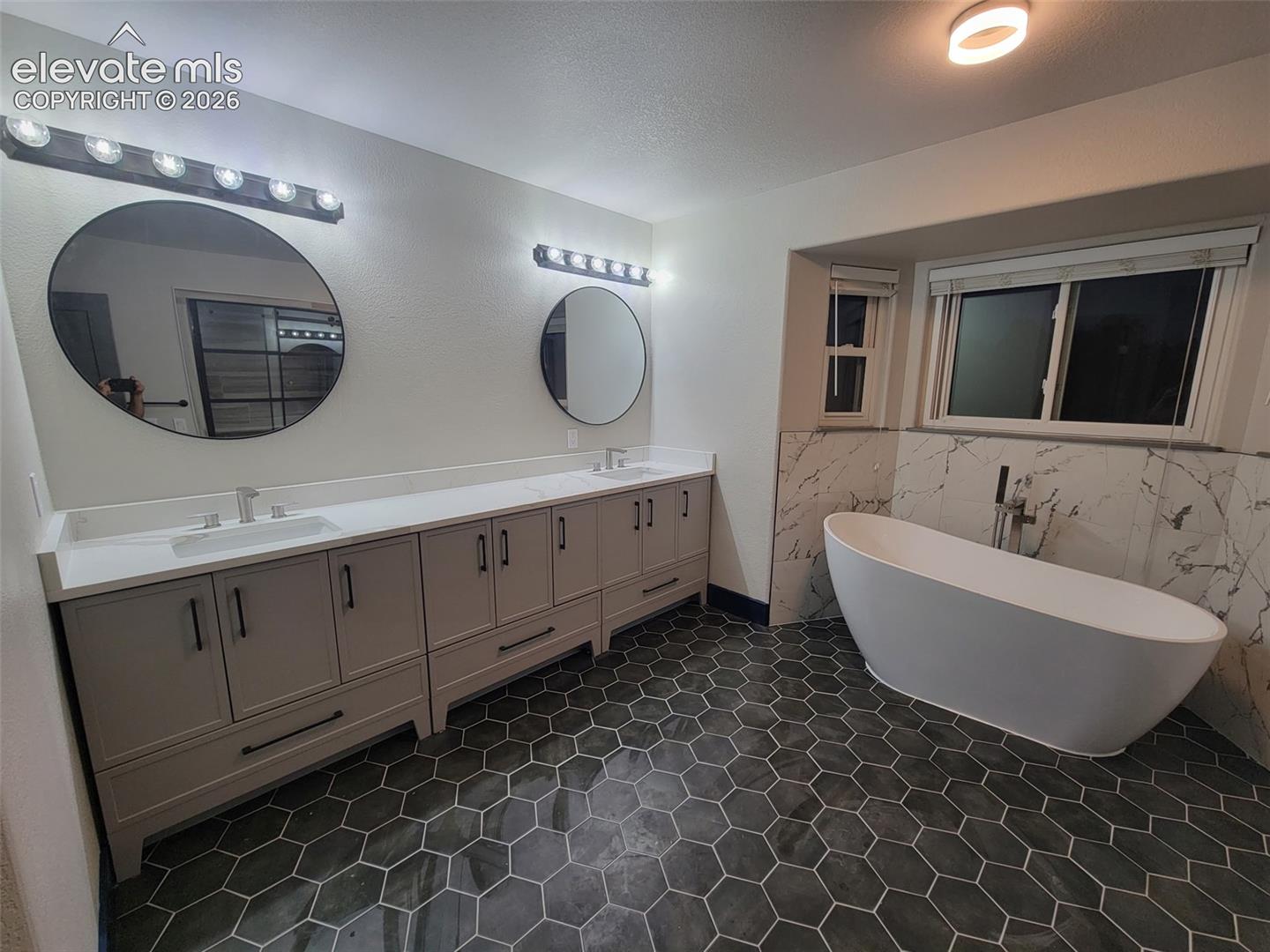 Freestanding soaking tub, dual vanity with undermount sinks, hexagon tile flooring, and wall-mounted light fixtures