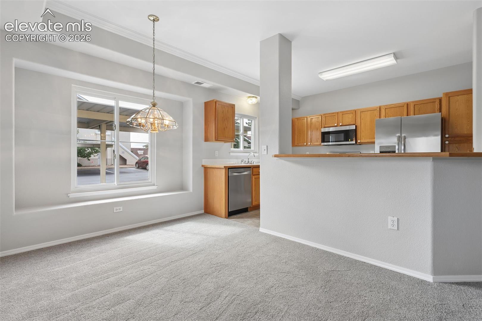 Recessed window with deep sill, overhead chandelier, light wood-finish cabinetry, stainless steel appliances, and gray carpet flooring