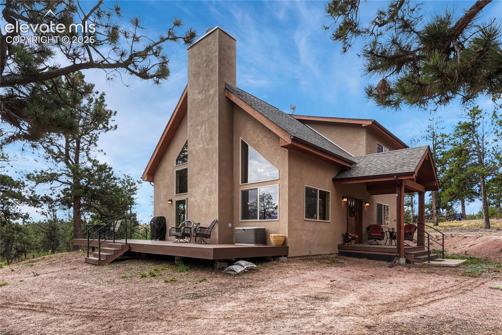 Stucco exterior featuring a prominent chimney, angular geometric windows, a wrap-around wooden deck, and a covered front porch