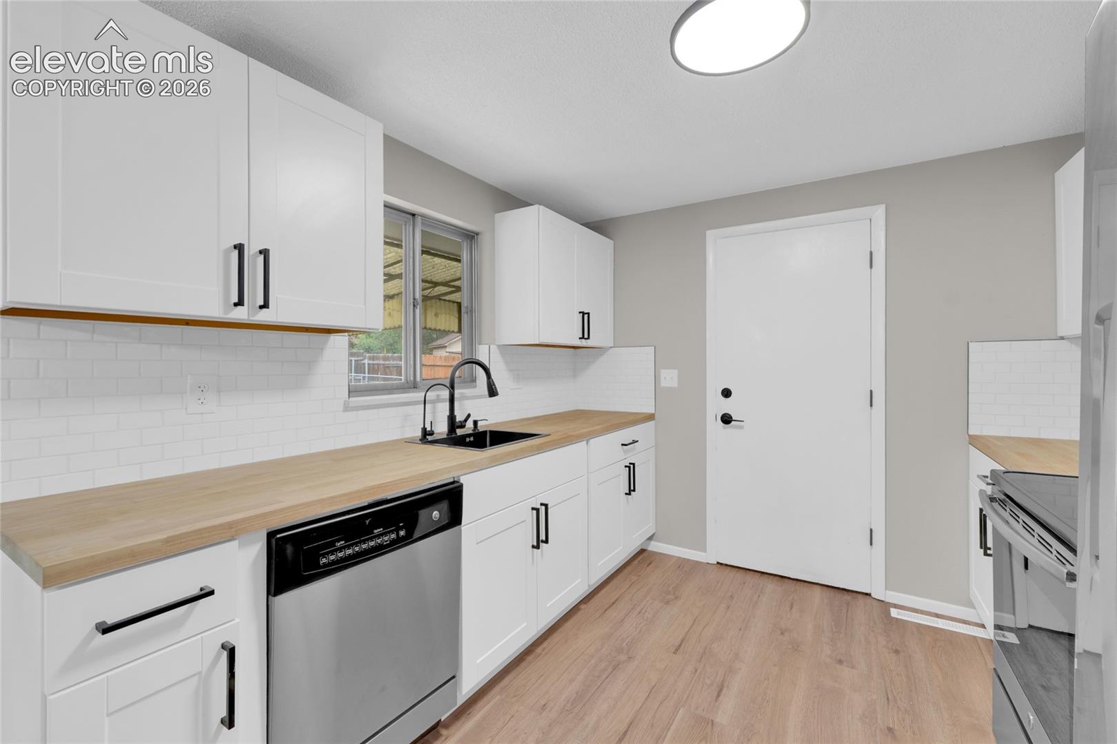 White shaker-style cabinetry with matte black hardware, wood-finish countertops, subway tile backsplash, stainless steel dishwasher, and wood-finish flooring