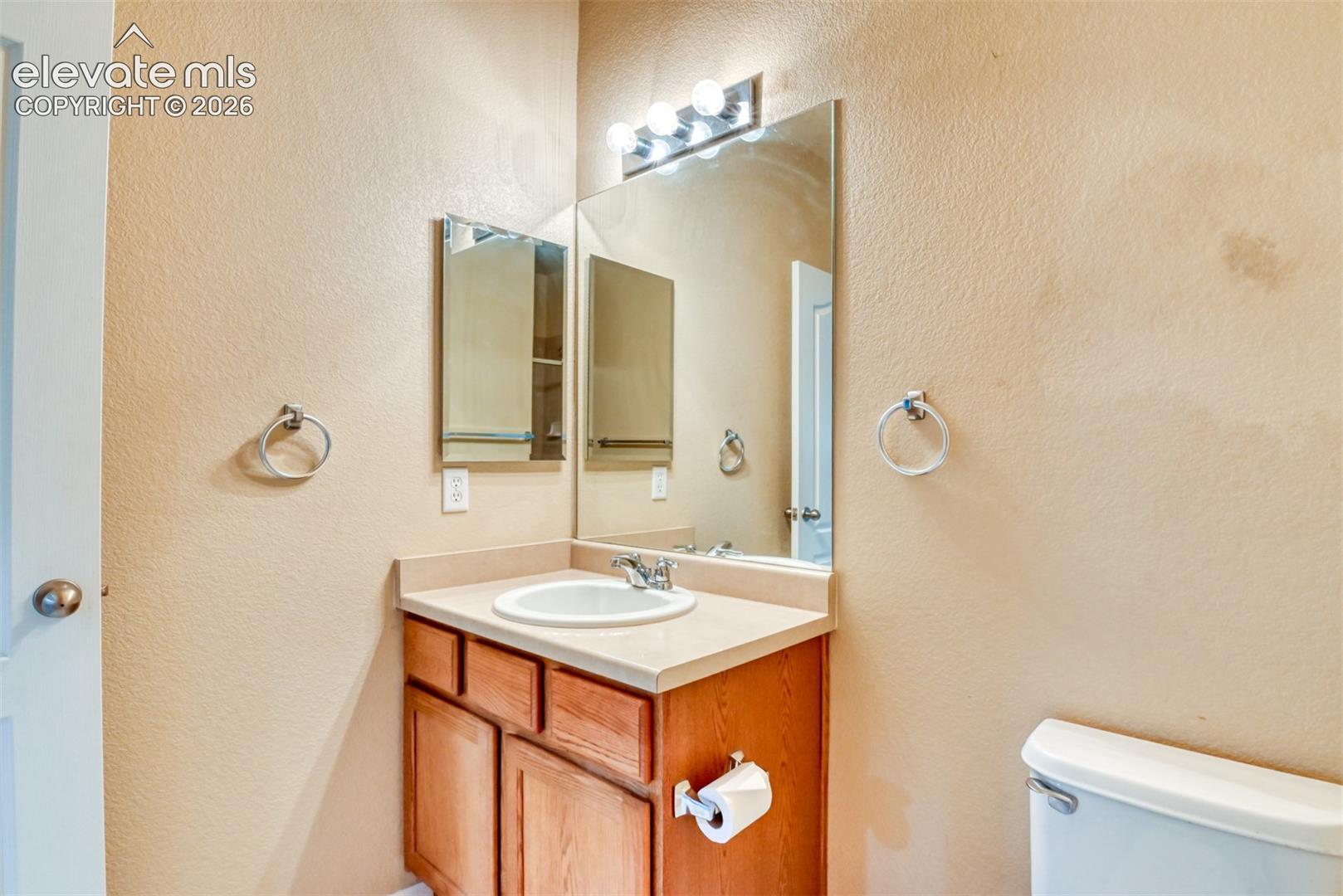 Bathroom vanity with wood-finish cabinetry, solid-surface countertop, integrated basin, wall-mounted mirror, and multi-bulb vanity lighting