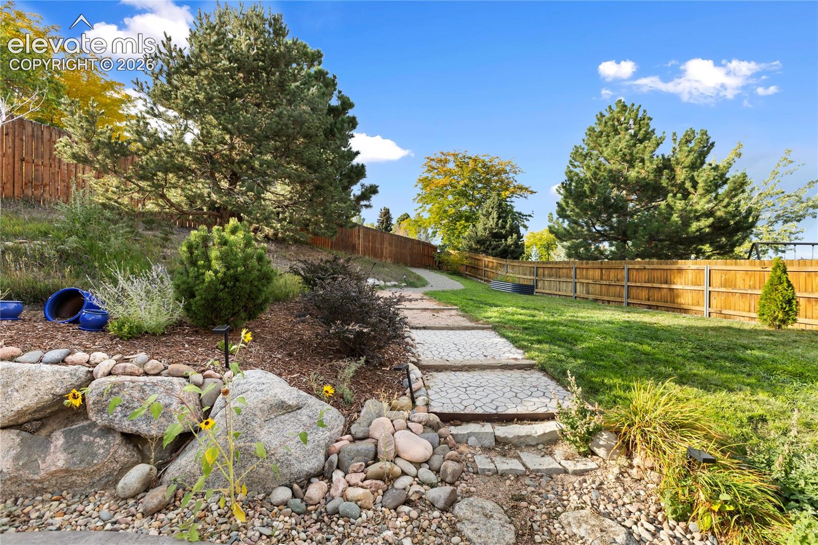 Expansive backyard featuring a manicured lawn, established landscaping, a stone retaining wall, a paved pathway, and a wood privacy fence
