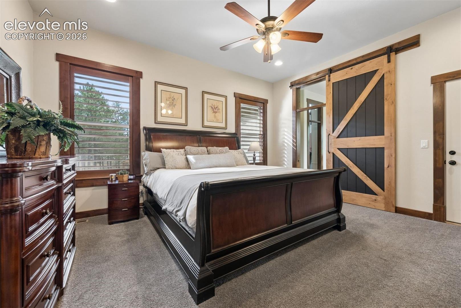 Natural wood-trimmed windows, neutral carpeting, a ceiling fan with integrated lighting, and a sliding barn door featuring a wood frame and dark paneling
