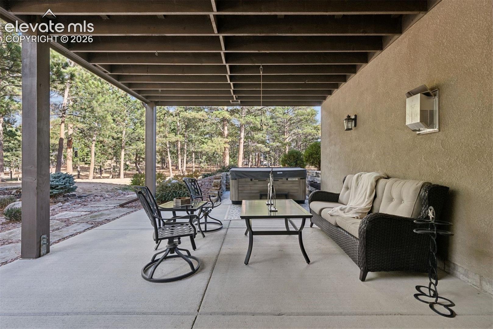 Covered concrete patio featuring an exposed-beam ceiling, stucco exterior wall, and wall-mounted exterior lighting fixtures
