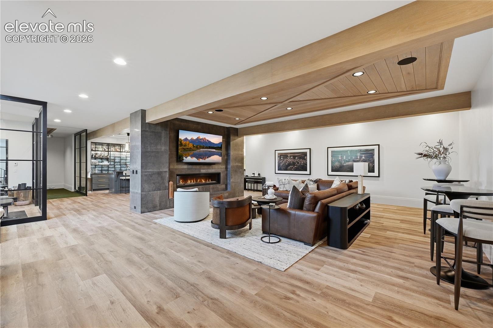 Wood-finish flooring, custom wood-paneled ceiling inlay with recessed lighting, linear fireplace with large-format stone surround, and floor-to-ceiling metal-framed glass partition