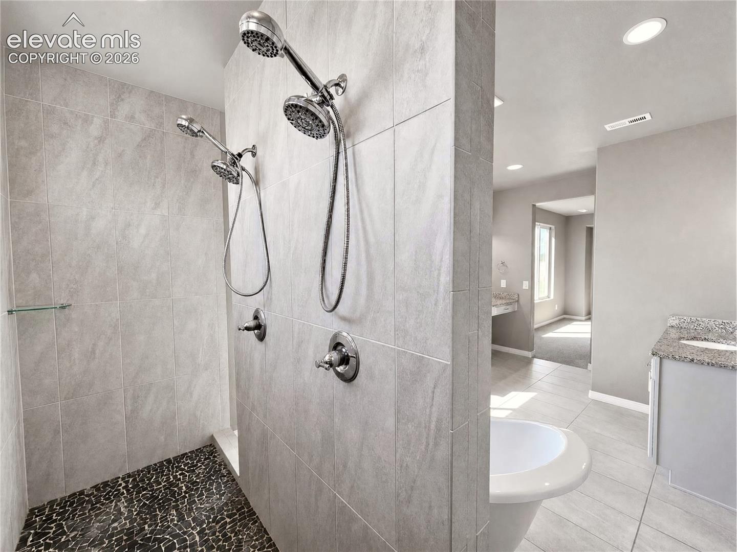 Walk-in shower with dual shower heads, light-toned tile walls, and a pebble-finish floor