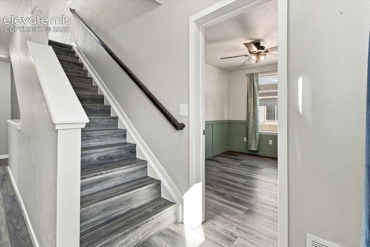 Wood-finish flooring throughout, a staircase with a dark wood handrail, a room with wainscoting, a window, and a ceiling fan