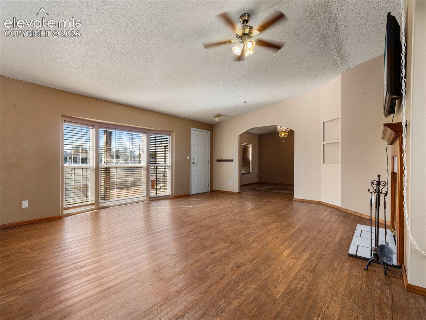 Interior featuring wood-finish flooring, a multi-pane window with blinds, a ceiling fan, an arched entryway, and a fireplace with a mantel