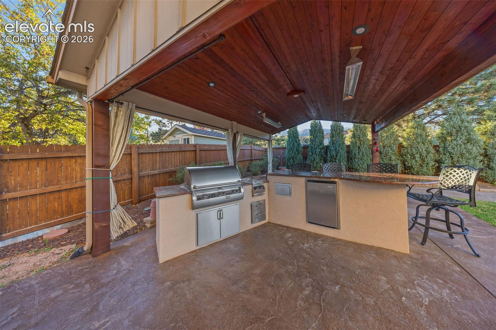 Covered outdoor kitchen featuring a wood-plank ceiling, stamped concrete flooring, built-in stainless steel grill, under-counter refrigeration, and stone-finish bar seating