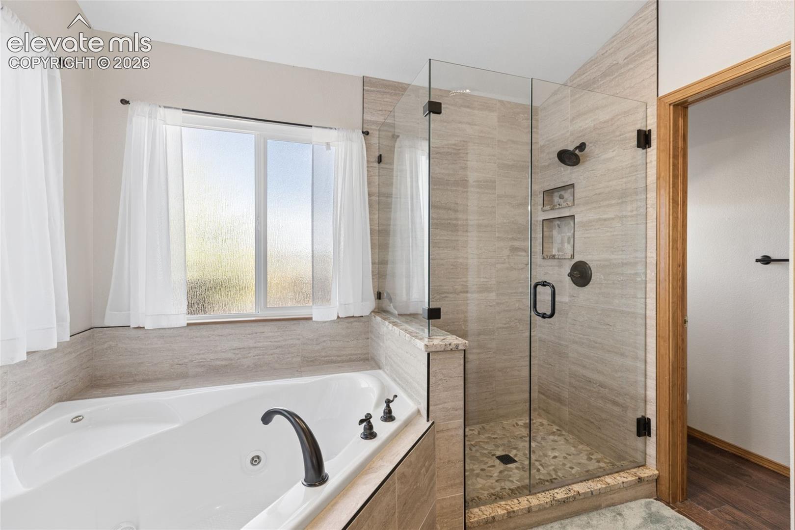 Jetted soaking tub with deck-mounted hardware, glass-enclosed walk-in shower with stone tile surround, stone-topped half wall, pebble-style shower floor tiling, and dark-finish plumbing fixtures