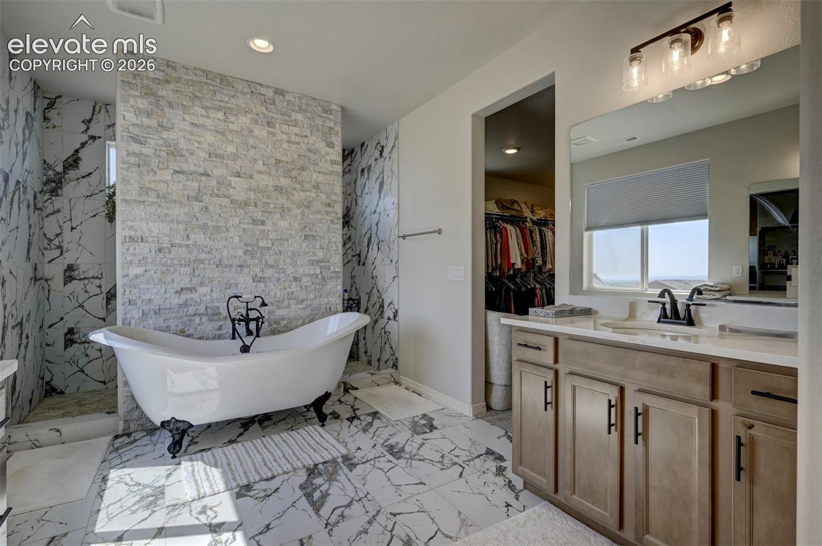 Bathroom featuring a freestanding clawfoot tub, floor-to-ceiling marbled tile, a stacked stone accent wall, and a wood-finish vanity with dual sinks