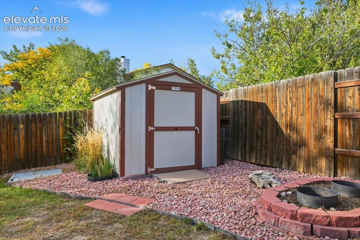 Freestanding storage shed with wood-finish siding, positioned on a red gravel bed, bordered by a wooden privacy fence, and featuring a brick-lined fire pit