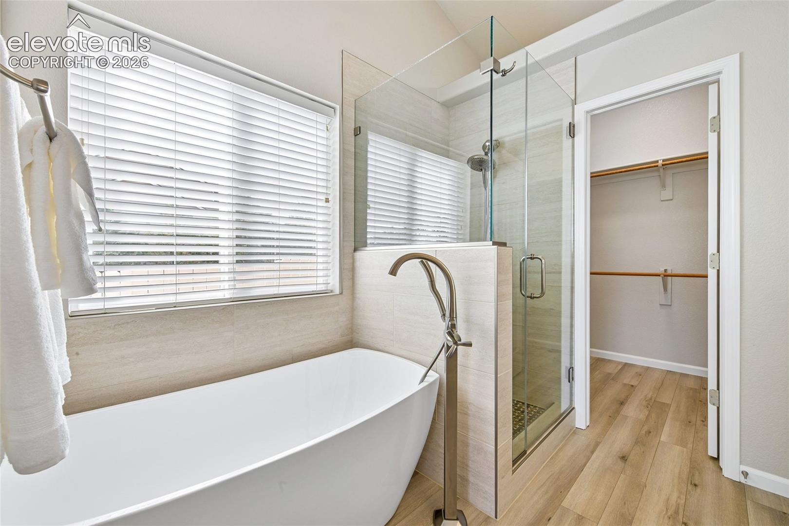 Freestanding soaking tub with floor-mounted faucet, glass-enclosed shower, wood-finish flooring, and a walk-in closet