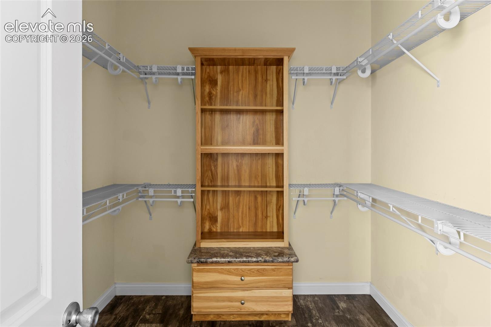 Walk-in closet featuring wood-finish flooring, built-in wooden cabinetry with open shelving, two storage drawers, and a stone-pattern countertop, complemented by perimeter wire shelving