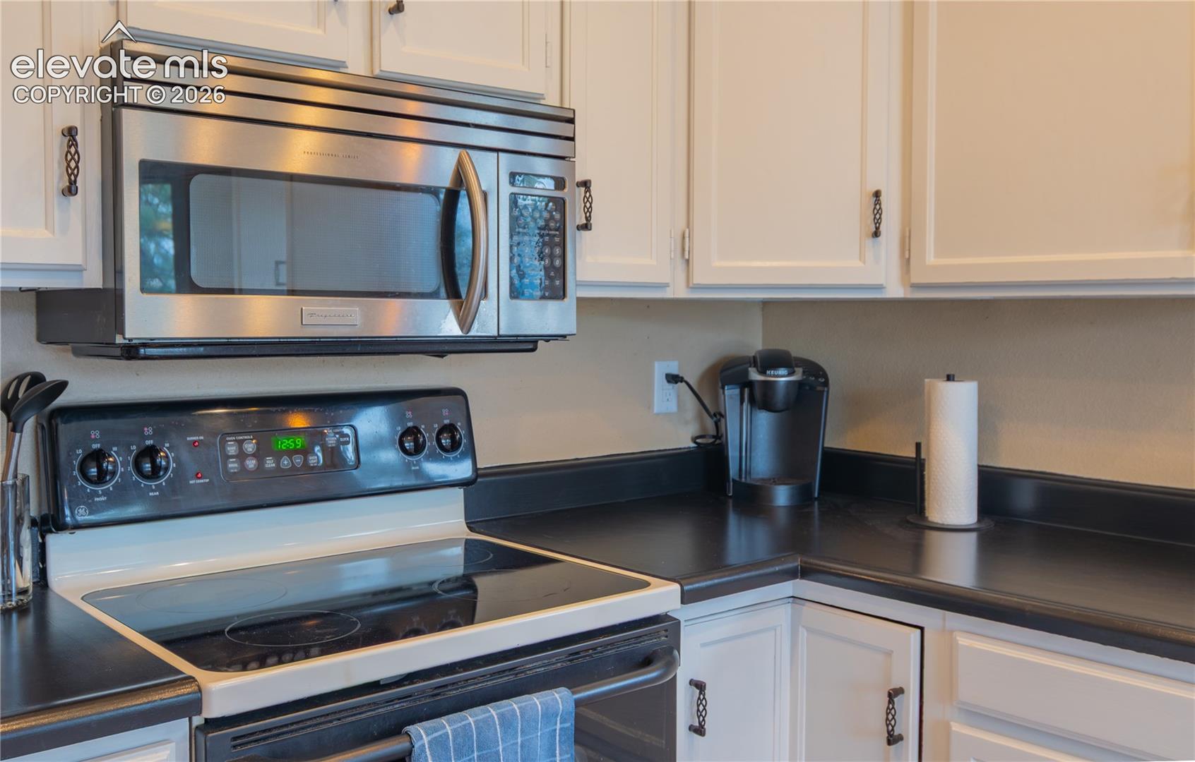 White cabinetry with dark ornamental hardware, black countertops, stainless steel over-the-range microwave, and a black electric range