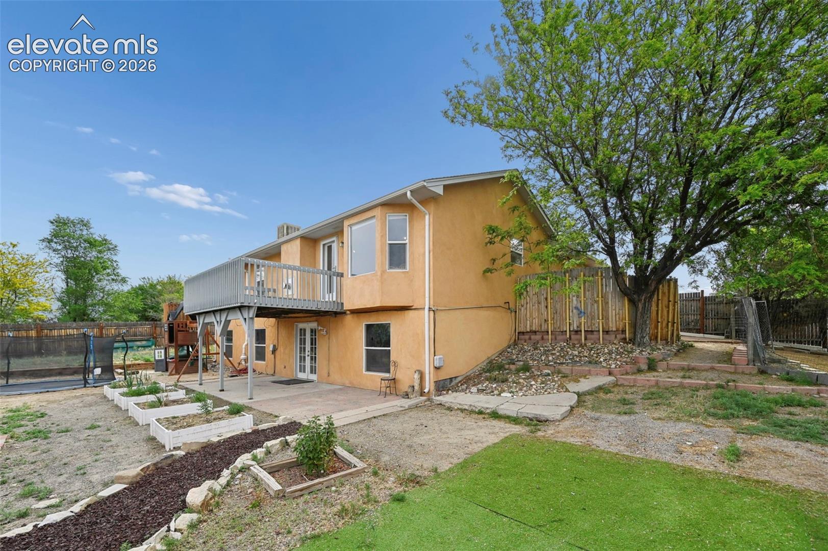 Rear exterior featuring a second-story deck, ground-level patio, multiple raised garden beds, a large mature tree, and artificial turf