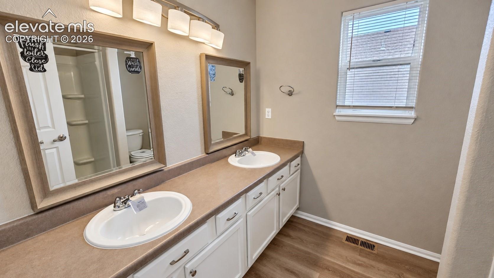 Dual-vanity bathroom featuring white cabinetry, solid-surface countertops with integrated dual sinks, wood-finish flooring, and wall-mounted light fixtures