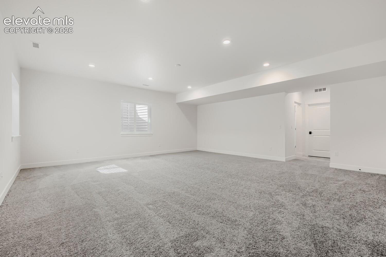 Expansive room featuring plush gray carpeting, recessed ceiling lighting, a window with plantation shutters, and a white paneled door