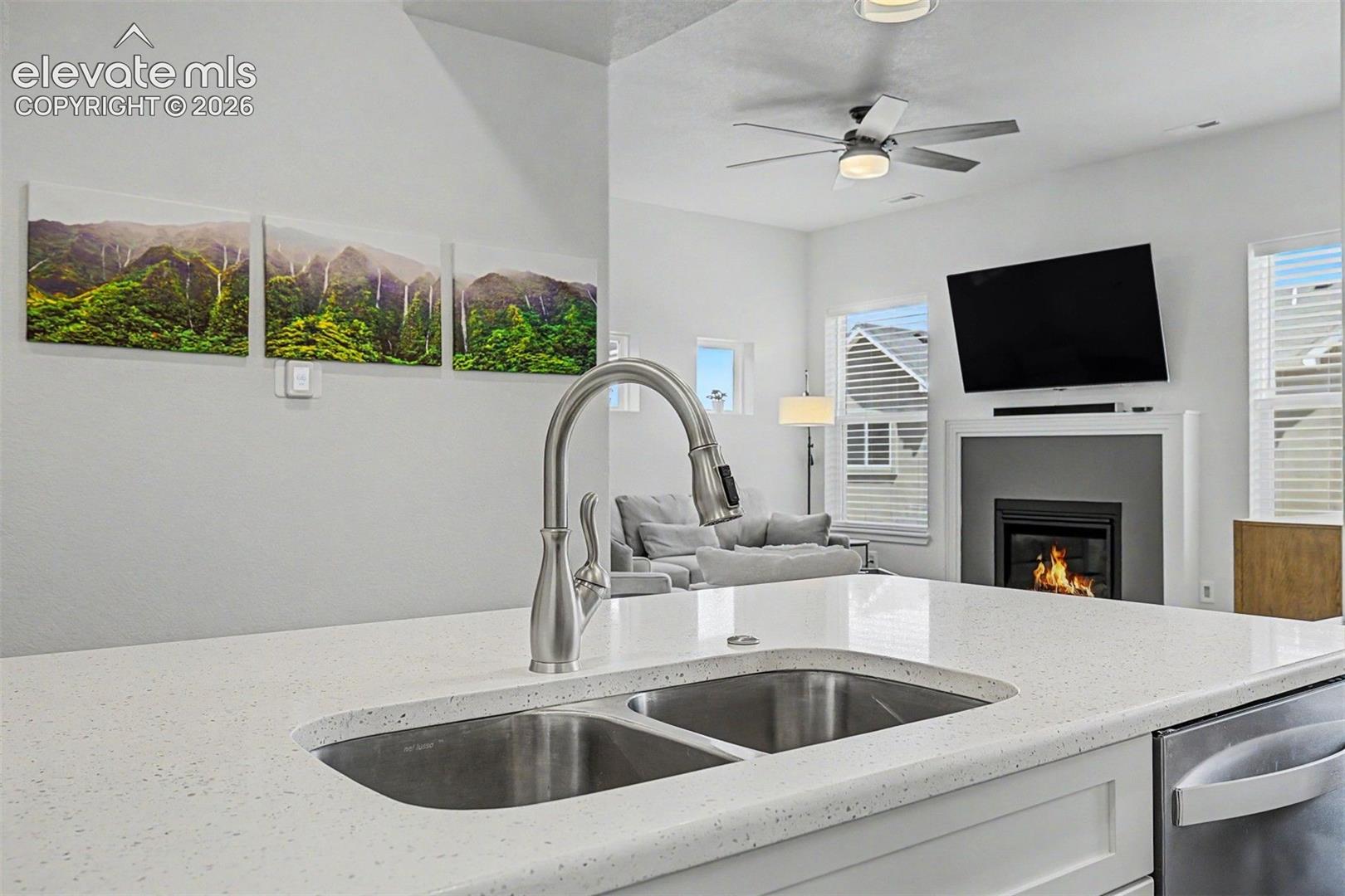 At the island sink, the view extends all the way into the living room, where the fireplace flickers and lush nature-inspired artwork lines the walls — proof that even the most everyday moments in this home come with a beautiful backdrop.