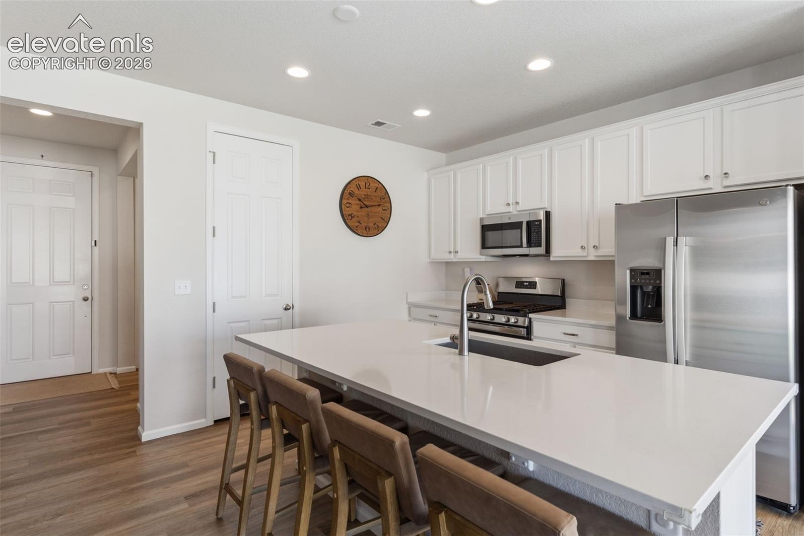 Modern kitchen featuring a large central island with seating, white shaker-style cabinetry, stainless steel appliances, recessed lighting, and wood-finish flooring