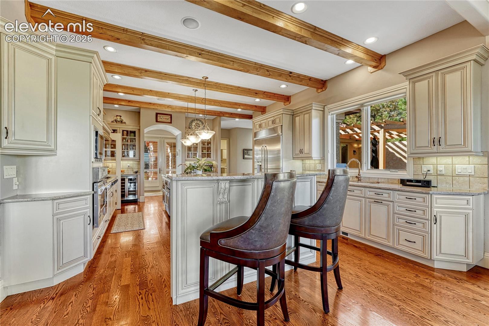 Kitchen featuring light-toned cabinetry, natural stone countertops, wood-finish flooring, exposed ceiling beams, and integrated stainless steel appliances