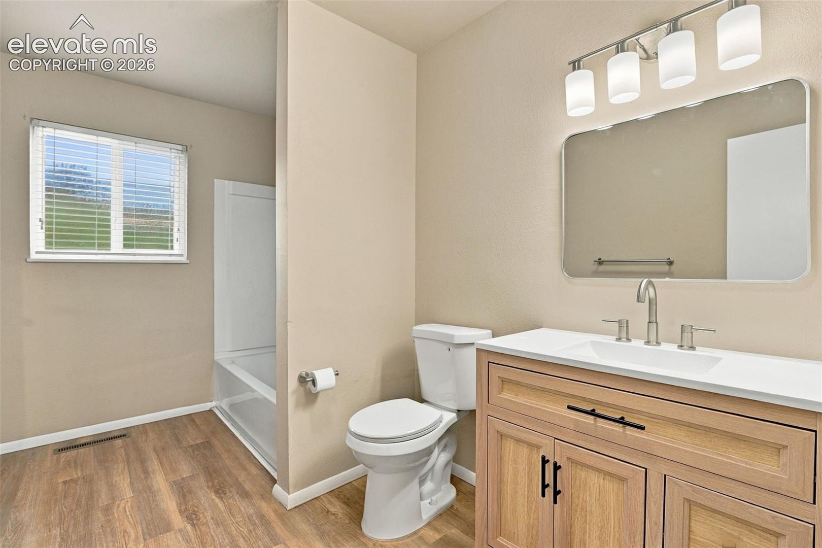 Bathroom featuring a wood-finish vanity with a light-toned countertop, rectangular wall mirror, four-light vanity fixture, and wood-finish flooring