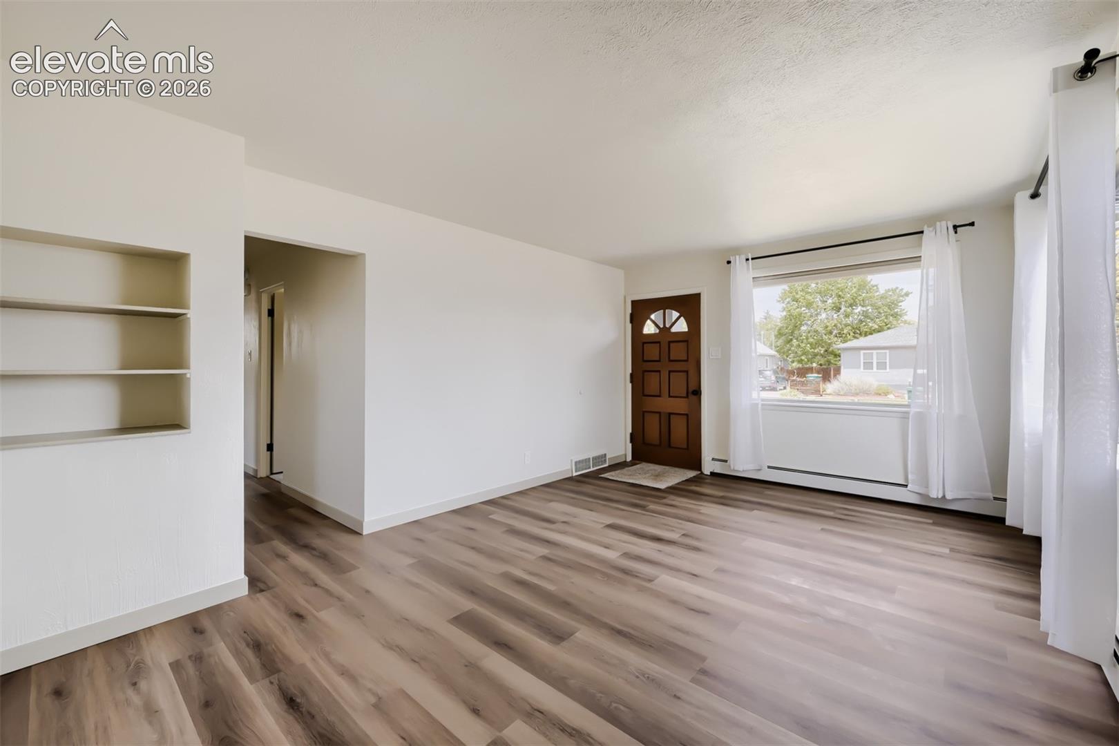 Spacious living area showcasing wood-finish flooring throughout, abundant natural light from multiple windows and glass doors, built-in shelving, and a decorative wooden entry door with architectural detailing