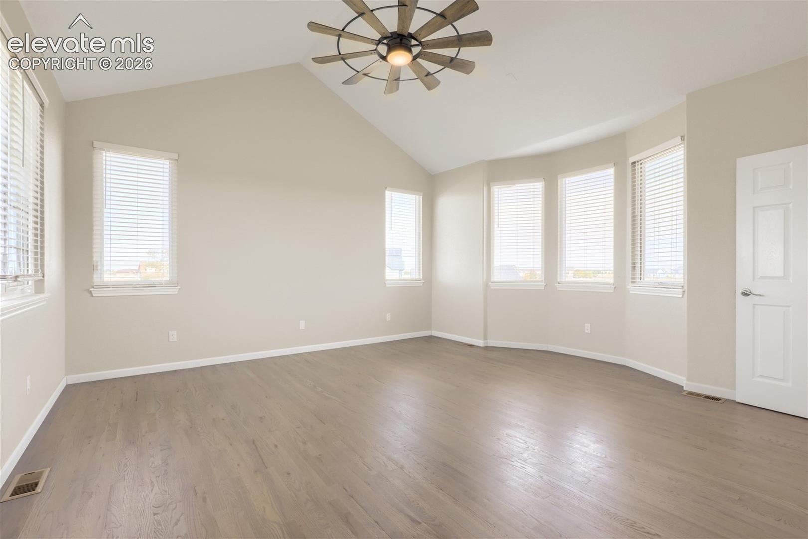 Spacious and serene main floor primary suite featuring dramatic vaulted ceilings and a statement windmill ceiling fan. Beautiful hardwood floors sweep through the room, while a sun-drenched bay of large windows floods the space with natural light. Facing west, this room captures breathtaking mountain views and golden hour sunsets. Finished in a warm neutral palette, this generous retreat offers a peaceful, airy sanctuary.