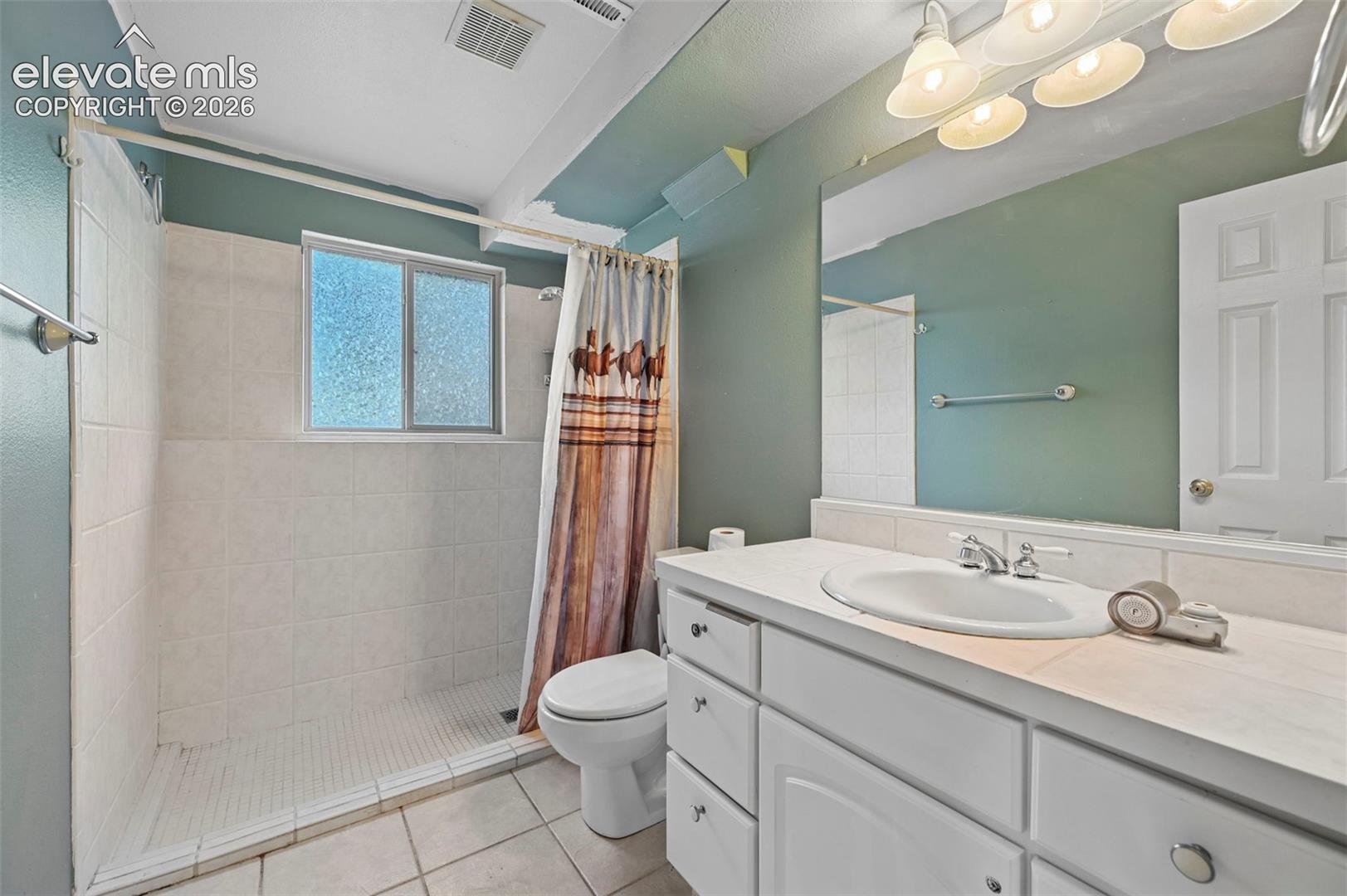 Bathroom with white tile flooring, a walk-in shower featuring white tile walls, a double-pane window, and a white vanity with an integrated sink