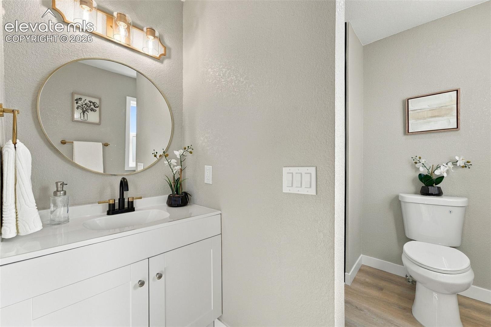 Modern vanity featuring a white cabinet, stone-finish countertop with an integrated basin, matte black faucet, oval mirror, and three-light wall-mounted fixture, complemented by wood-finish flooring