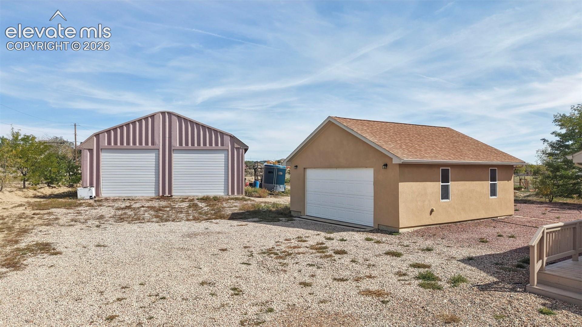 Detached structures including a metal two-bay garage and a single-bay stucco garage with a shingled roof, set on a gravel lot
