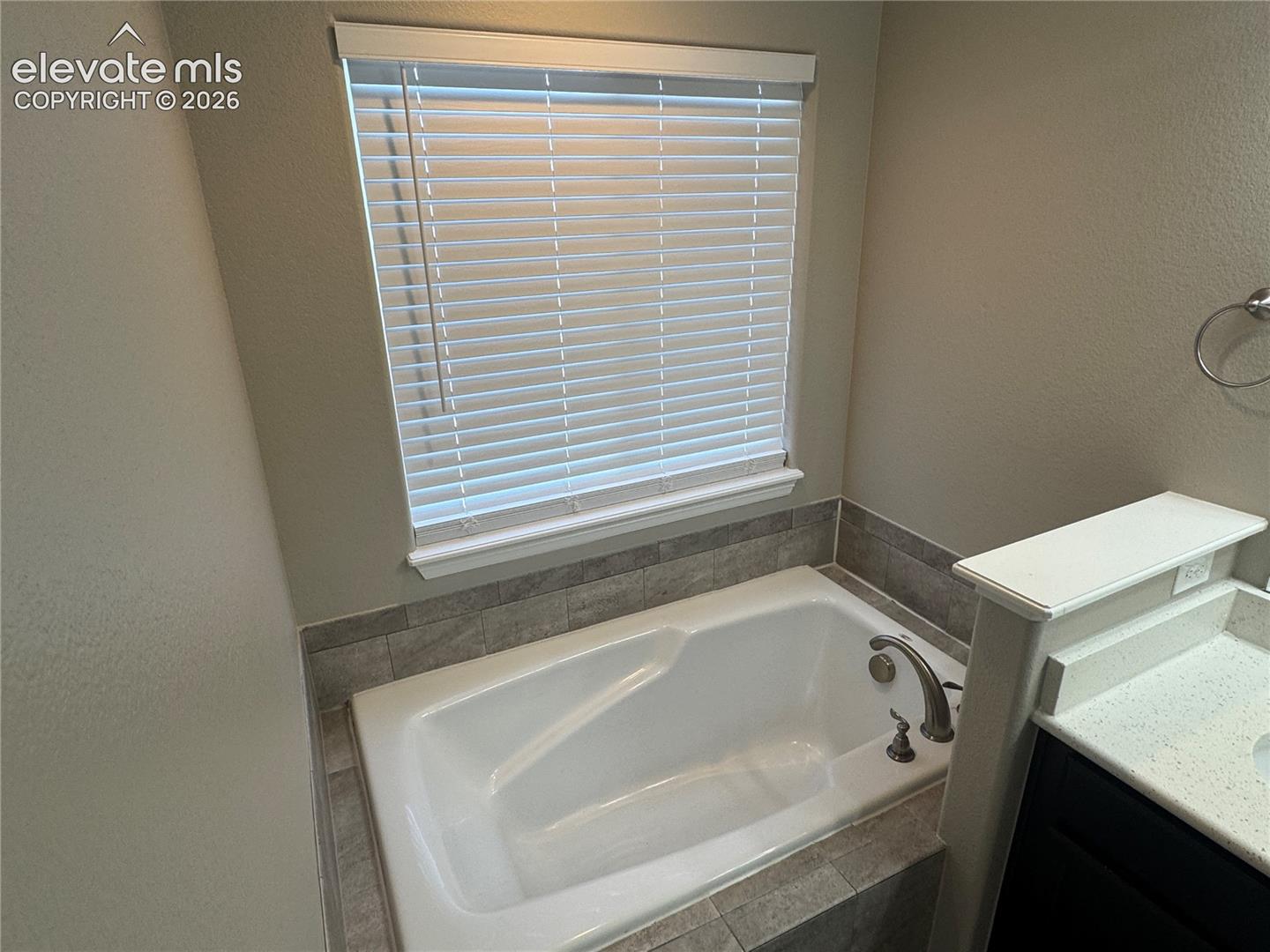 Built-in soaking tub with a tiled surround, neutral wall finish, installed window blinds, stone-finish vanity countertop, and a wall-mounted metal towel ring