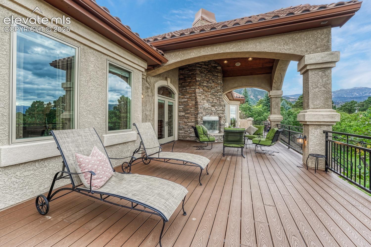 Covered outdoor deck featuring composite decking, a stone-faced fireplace, stucco columns, and a mountain backdrop