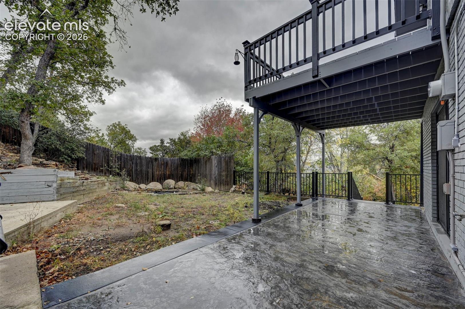 Covered concrete patio featuring a tiered wood-plank deck, black metal railings, and a fenced rear perimeter with stone retaining walls