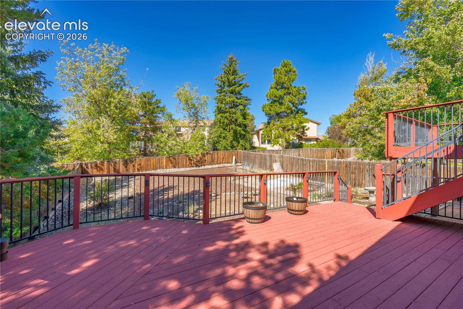 Expansive outdoor deck with red-stained wood flooring and black metal railings, featuring integrated stairs to the ground level and a surrounding wooden fence