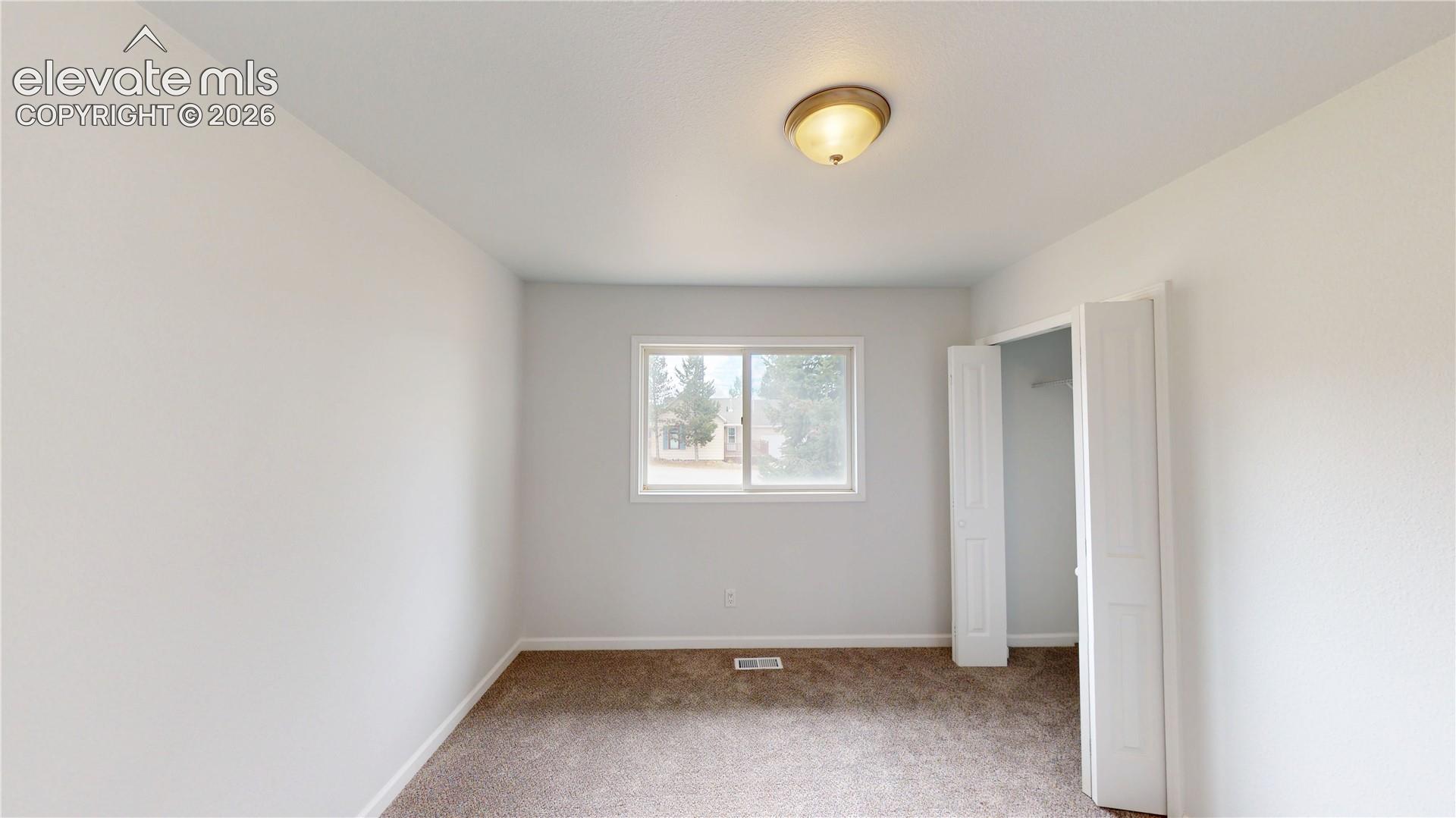 Neutral wall tones, carpeted flooring, a ceiling-mounted light fixture, a window with white trim, and a bi-fold closet door