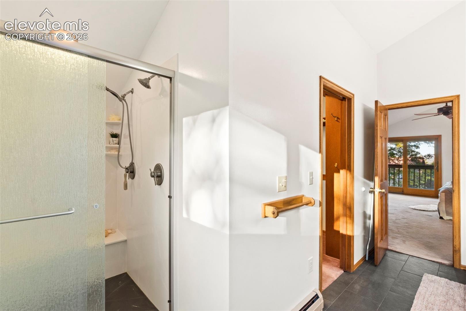 Shower enclosure with glass door, handheld shower head, and built-in bench seating, featuring dark tile flooring and a natural wood-finish door frame