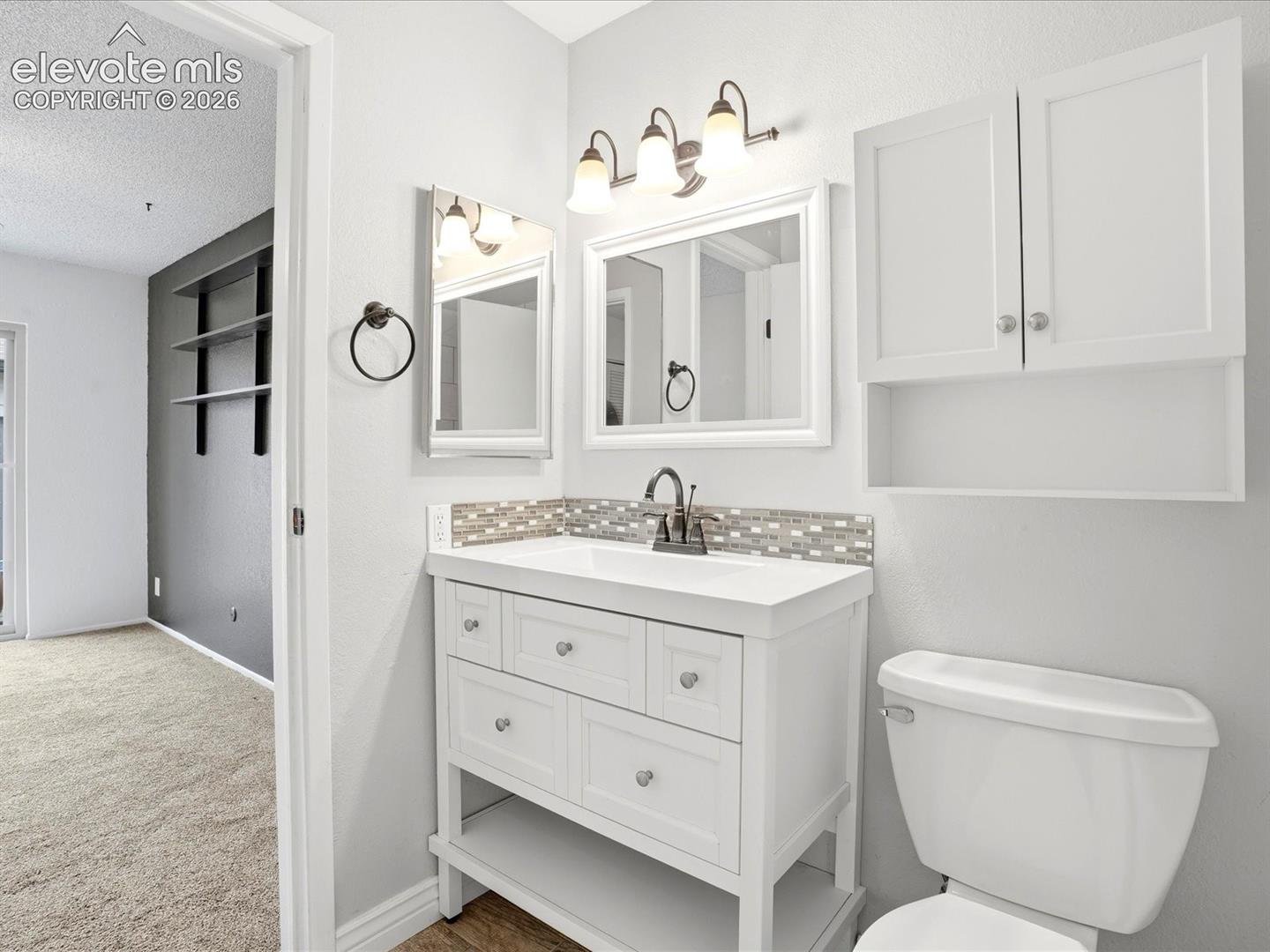 White vanity with an integrated sink, mosaic tile backsplash, brushed metal hardware, three-light vanity fixture, and white wall-mounted storage cabinetry