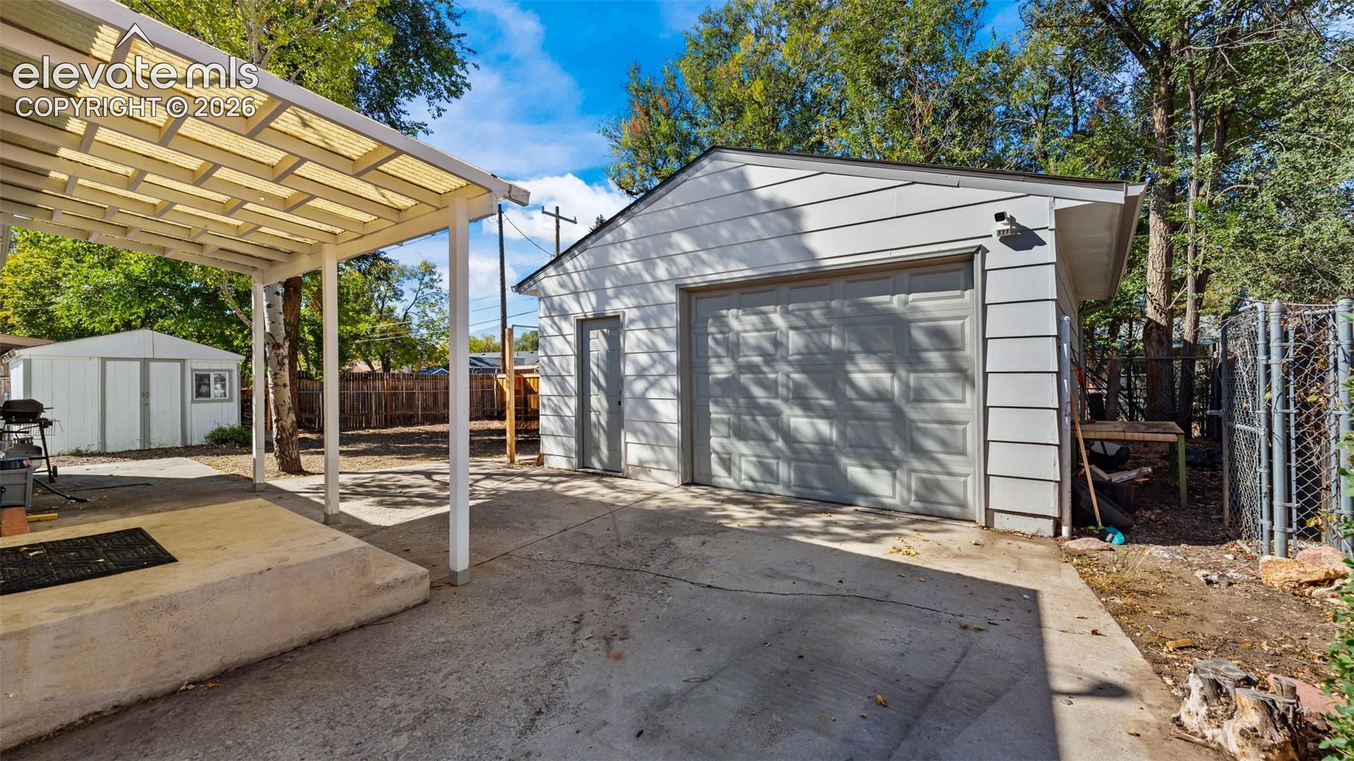 Detached garage with horizontal siding, covered patio area with a semi-transparent roof, concrete slab, and supplemental storage shed