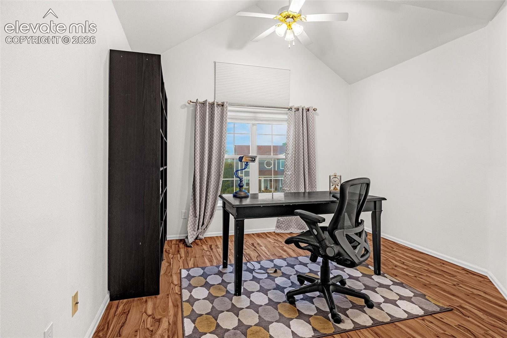 Vaulted ceiling with a ceiling fan and light fixture, wood-finish flooring, and a large window with privacy blinds