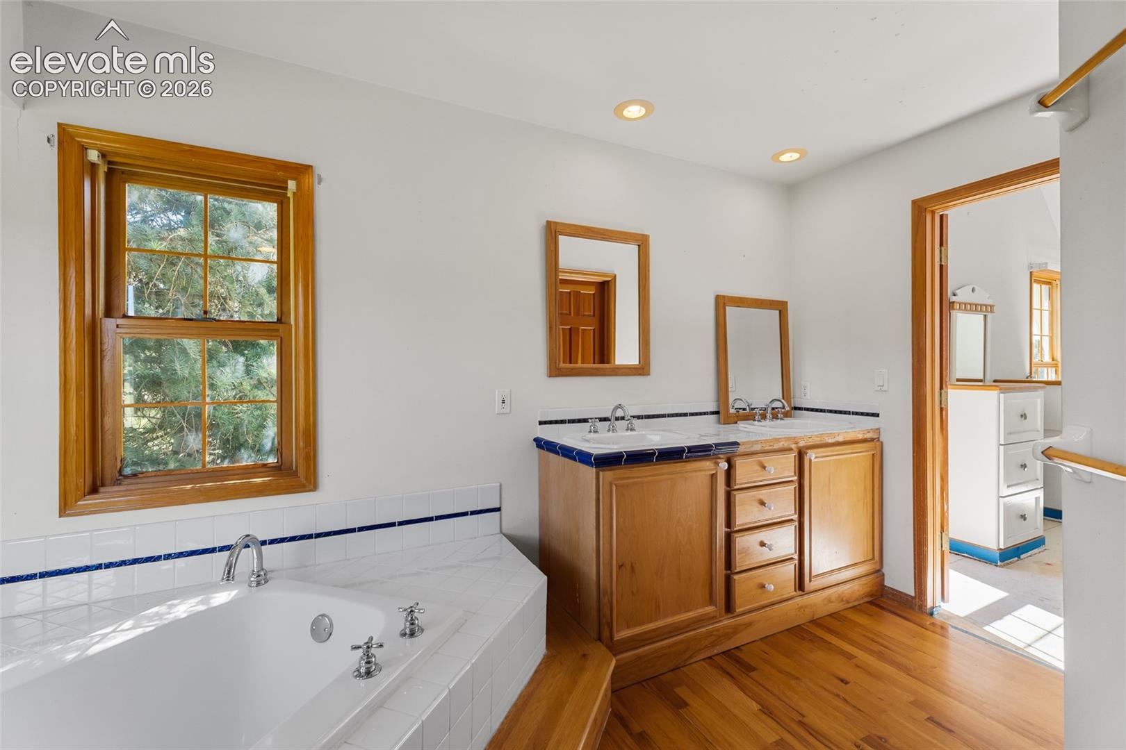 Corner soaking tub with tiled surround, wood-frame window, dual vanity with wood cabinetry, and recessed lighting
