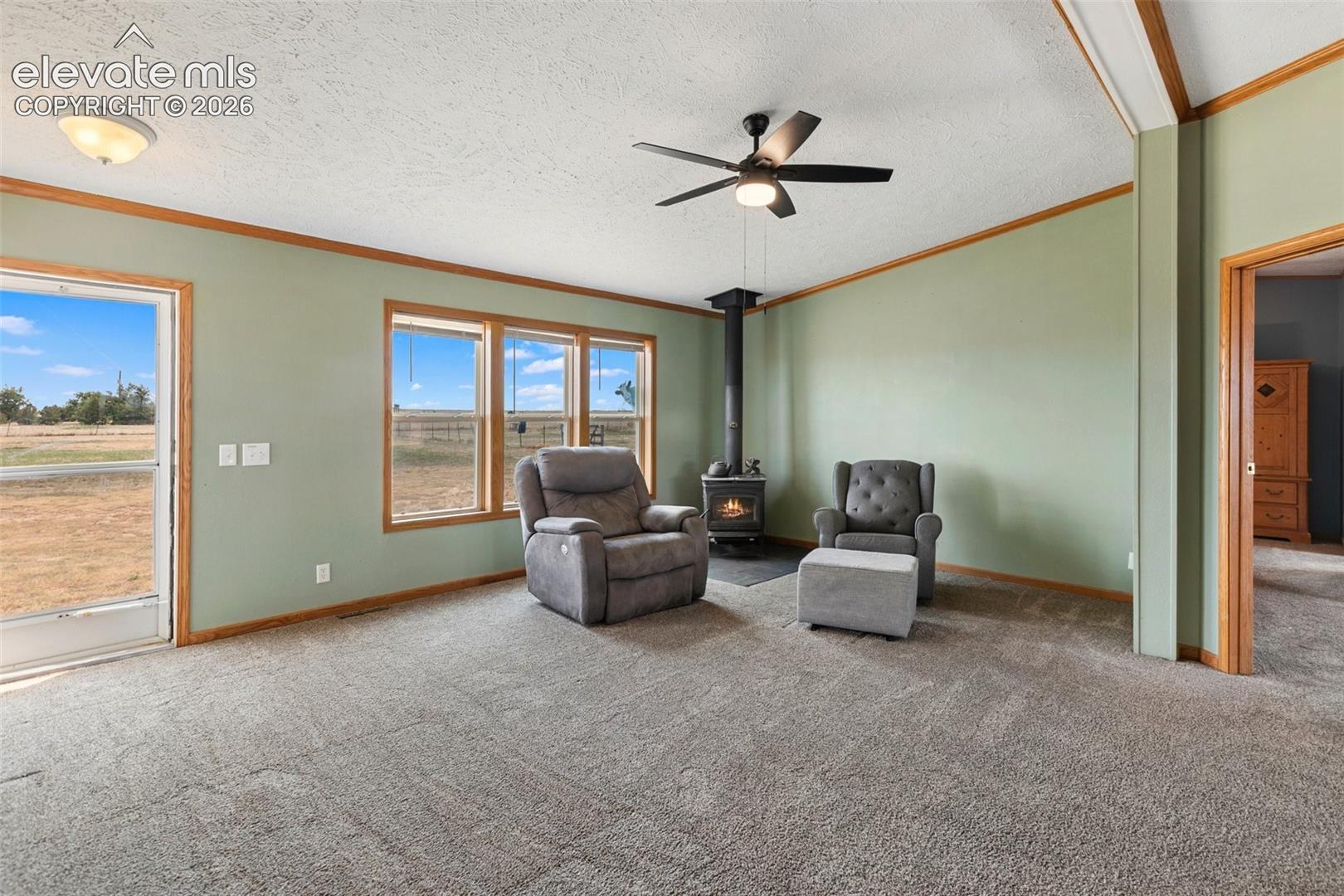 Living area featuring a freestanding wood-burning stove, three large windows with wood trim, a glass-paneled exterior door, and a ceiling fan with integrated lighting