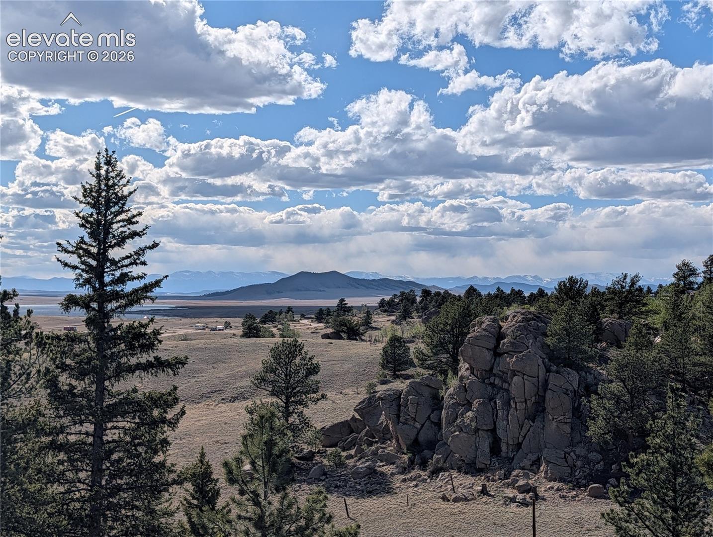Expansive mountain landscape featuring prominent rock formations, mature evergreen trees, and a distant body of water