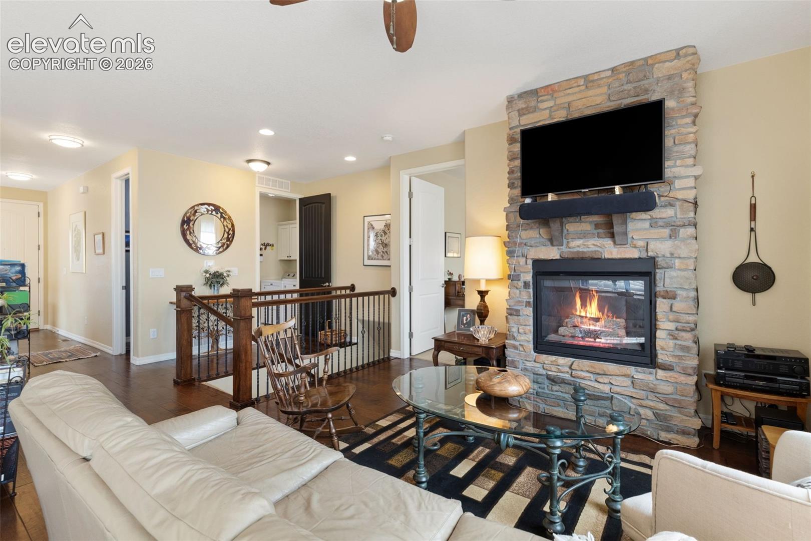 Living area featuring a stone-clad fireplace with a hearth, dark wood-finish flooring, a wood staircase with wrought iron balusters, and recessed ceiling lighting.
